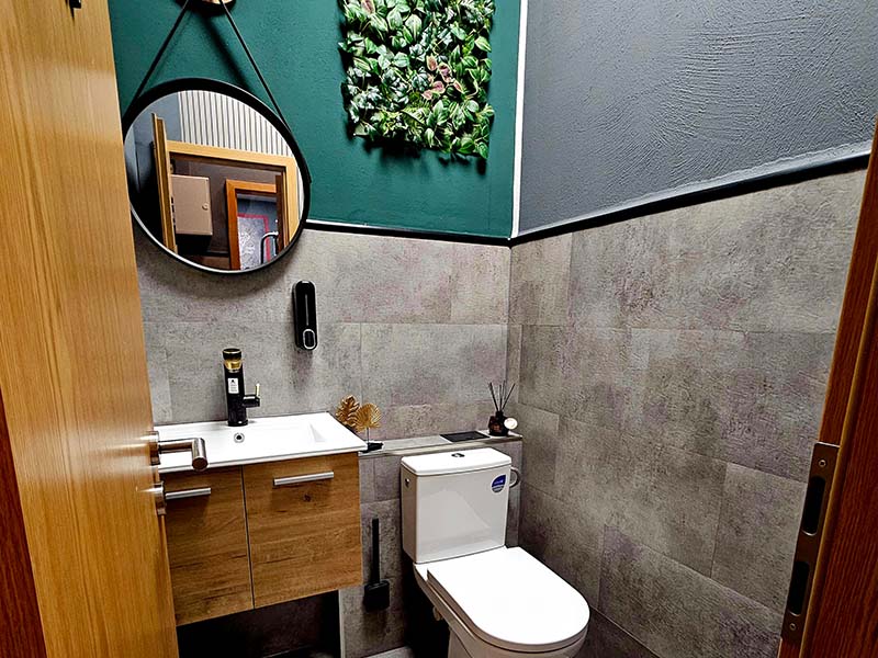 Hochwertiges Badezimmer im TWENTY FOUR SEVEN fit Beuna, Merseburg Modernes Badezimmer im Fitnessstudio TWENTY FOUR SEVEN fit in Beuna, Merseburg, mit stilvollem Holz-Waschtisch, rundem schwarzem Spiegel und grauen Fliesen.