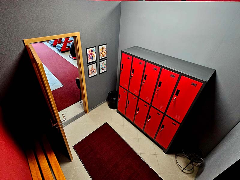 Umkleidekabine – TWENTY FOUR SEVEN fit Beuna, Merseburg Moderne Umkleidekabine mit roten Spinden, grauen Wänden und Holzbank im Fitnessstudio TWENTY FOUR SEVEN fit in Beuna, Merseburg.